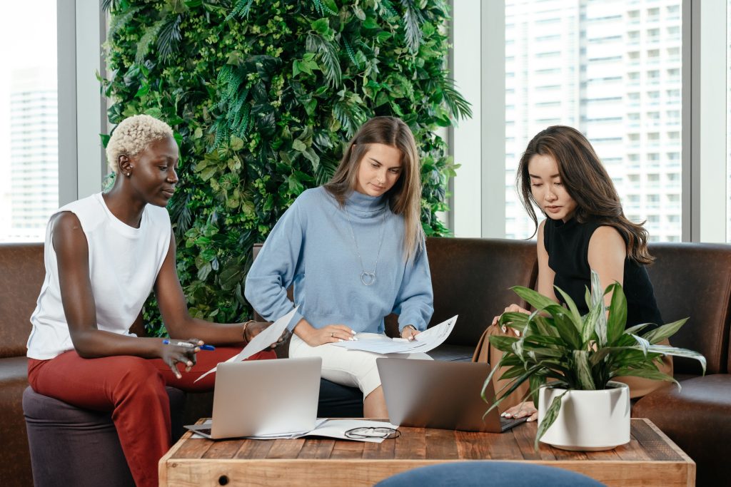 ujeres profesionales colaborando en el trabajo, reflejando liderazgo femenino y bienestar laboralFocused multiethnic female coworkers with documents sitting at wooden desk with notebooks and flowerpot while working together in modern workspace